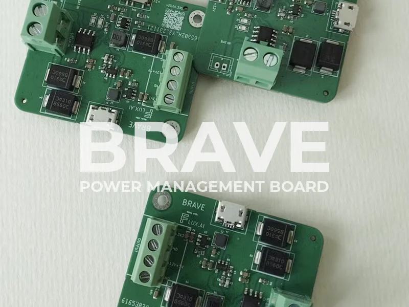 This image features a close-up view of a green printed circuit board (PCB) labeled as a BRAVE POWER MANAGEMENT BOARD. The PCB is populated with various electronic components including integrated circuits (ICs), capacitors, resistors, and terminal blocks for connections. Some of the ICs have visible labels, and there are white printed labels and codes that indicate the board's layout and specifications. The text BRAVE is prominently displayed in a large white font at the center of the image, possibly serving as the brand or product name. The image seems to be meant for showcasing and highlighting its function in power management.